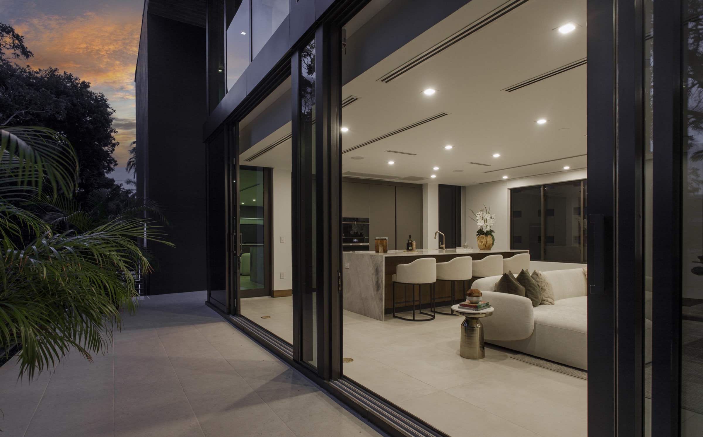 Completed Fair Isle Residence opening from warm interior spaces to its shaded pool terrace and tropical landscape at evening.
